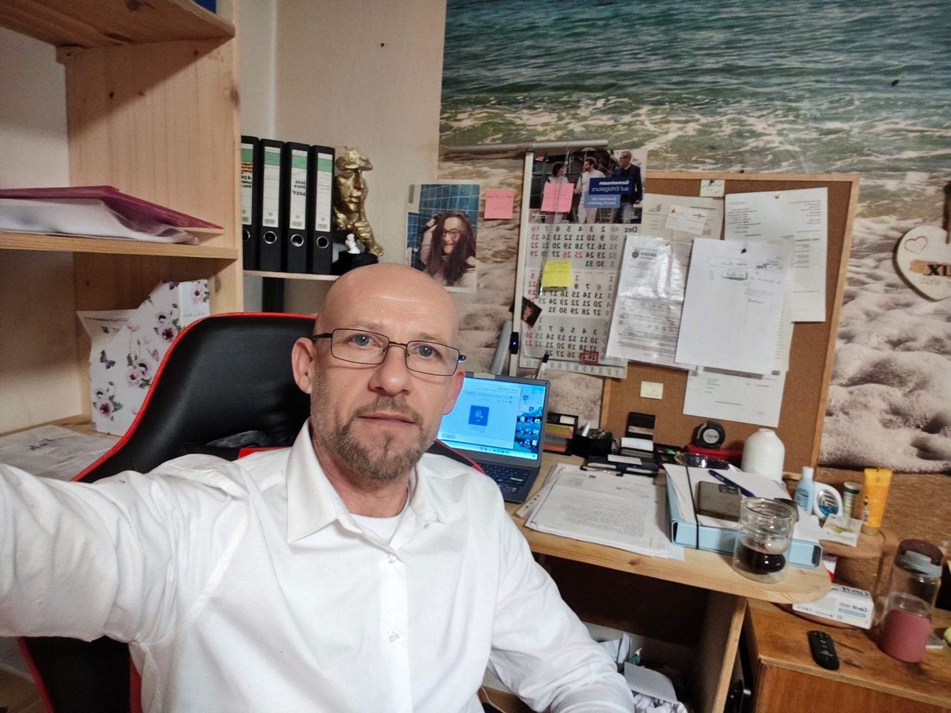 Man in white shirt and glasses sitting at desk in office with beach photo backdrop and organizational materials on cork board