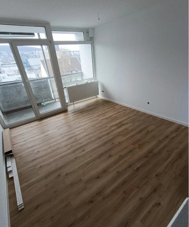 Empty apartment room with wooden flooring, white walls, and large windows overlooking a city skyline