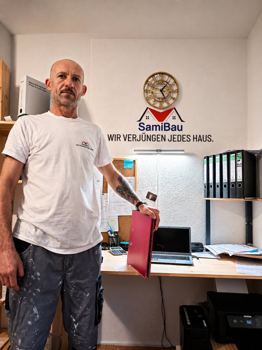 Man in white SamiBau t-shirt standing in office holding red folder, with company logo and clock on wall behind desk