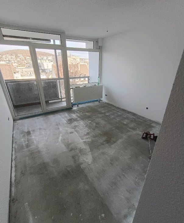 Empty apartment under construction with concrete flooring, white walls, and large glass balcony doors overlooking cityscape